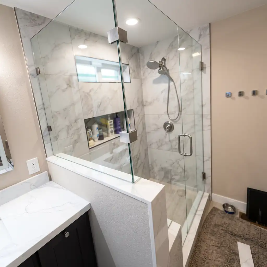modern minimalist bathroom interior featuring a walk-in shower enclosed by frameless glass panels with metallic hardware, surrounded by white marble-patterned tiles on walls and floor; adjacent to the shower is a vanity with a matching marble countertop and dark cabinetry, illuminated by recessed ceiling lights casting soft, even illumination; visible elements include a built-in niche shelf holding toiletries, a handheld showerhead with chrome fixtures, a pet food bowl on the floor near a black pet gate, and a mirror above the sink reflecting part of the room; camera angle is eye-level medium shot capturing the space from slightly off-center perspective, emphasizing clean lines and luxury materials