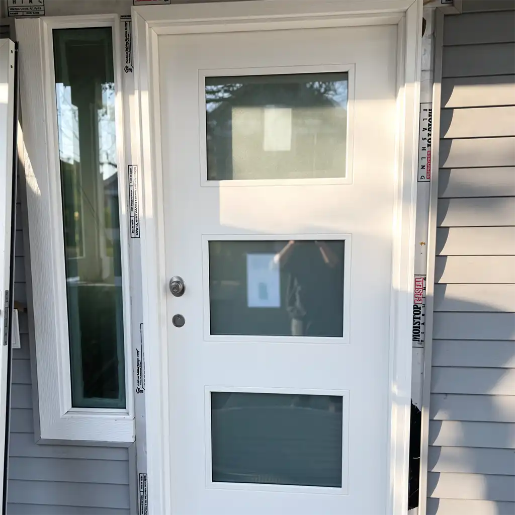 a modern white front door with three vertically aligned rectangular glass panels, framed by a matching white door jamb and flanked by white siding with horizontal slats; the door features a silver doorknob and deadbolt on the left side, with visible manufacturer labels on the door frame indicating “MOISTSTOP” and “FILLING” along the vertical edges; the scene is set outdoors during daytime under natural sunlight casting sharp shadows, with no human subjects present, emphasizing the architectural detail and clean lines of the door against a neutral gray exterior wall, captured in a medium shot from eye level with shallow depth of field isolating the door as the central focus