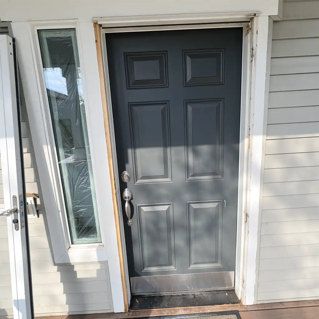 a modern residential front entrance featuring a dark matte gray six-panel security door with a brushed stainless steel handle and deadbolt, set within a white painted wooden frame showing signs of wear and minor damage along the edges; to the left, a tall narrow glass window with a white frame is partially removed or leaning against the wall, covered in protective plastic wrap, adjacent to a matching white-framed sliding glass door with a metallic handle visible on its side, all mounted on a light beige horizontal siding exterior; the scene is captured from a slightly low-angle eye-level perspective under bright natural daylight casting sharp shadows from nearby foliage onto the door’s surface, with a black rubber threshold mat placed at the base of the door and a small patch of weathered wooden deck flooring visible in the foreground