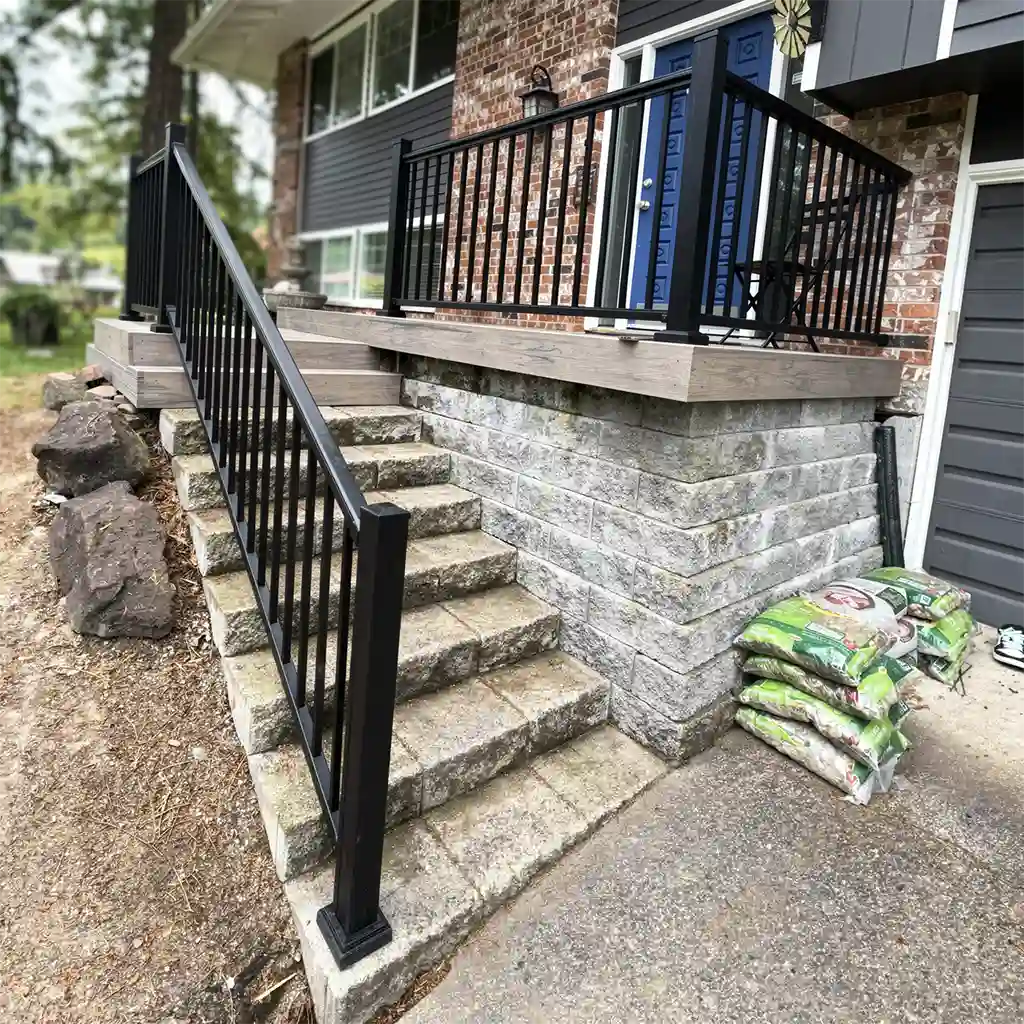 photorealistic outdoor residential scene featuring a modern house with dark gray siding and exposed brick facade, blue front door with decorative geometric panels, and a newly constructed wooden deck with black metal railings; wide stone staircase with matching black metal handrails leading up to the deck, flanked by stacked gray concrete retaining wall blocks and large natural boulders on the left side, with multiple green and white bags of soil or mulch piled near the base of the wall on the right, set against a backdrop of overcast daylight with soft diffused lighting, shallow depth of field emphasizing the staircase and railing details while blurring the background foliage and distant houses, low-angle perspective capturing the structure from ground level
