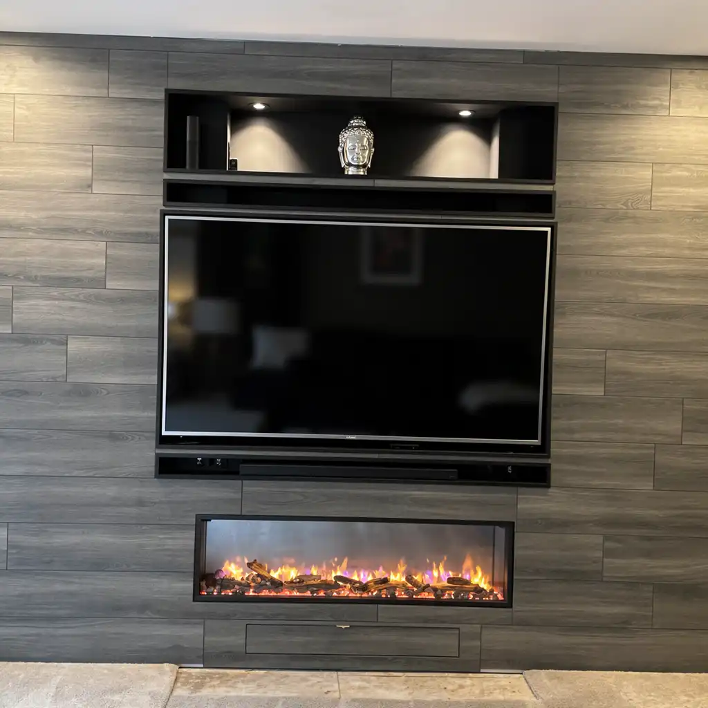 modern minimalist living room interior featuring a sleek, dark gray wood-paneled wall with horizontal plank texture, centered around a large flat-screen television mounted flush against the wall, flanked by a recessed black floating shelf above holding a silver Buddha head statue under two spotlights and a narrow black media console below; beneath the TV is an electric fireplace with realistic flickering flames in orange, yellow, and purple hues within a glass-enclosed rectangular frame, set into the wall with a matching dark gray console base; the floor is covered in light beige stone tiles, ceiling is white with no visible fixtures, ambient lighting is soft and warm from concealed sources creating subtle highlights on the wood grain and reflective surfaces, camera angle is eye level frontal shot capturing the entire wall unit
