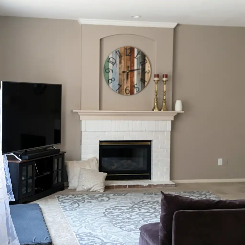 a modern living room interior featuring a white brick fireplace with a beige mantel, above which hangs a large circular rustic-style clock with multicolored wooden slats and black numerals, flanked by two tall gold candlesticks with red candles and a small white ceramic vase; to the left, a flat-screen television sits on a dark wood media console with glass-paneled doors, beneath it a light-colored patterned area rug covering part of the beige carpet, with a dark purple velvet sofa partially visible in the foreground, two cream-colored throw pillows resting beside the fireplace, walls painted in neutral taupe with white crown molding, ceiling recessed lighting