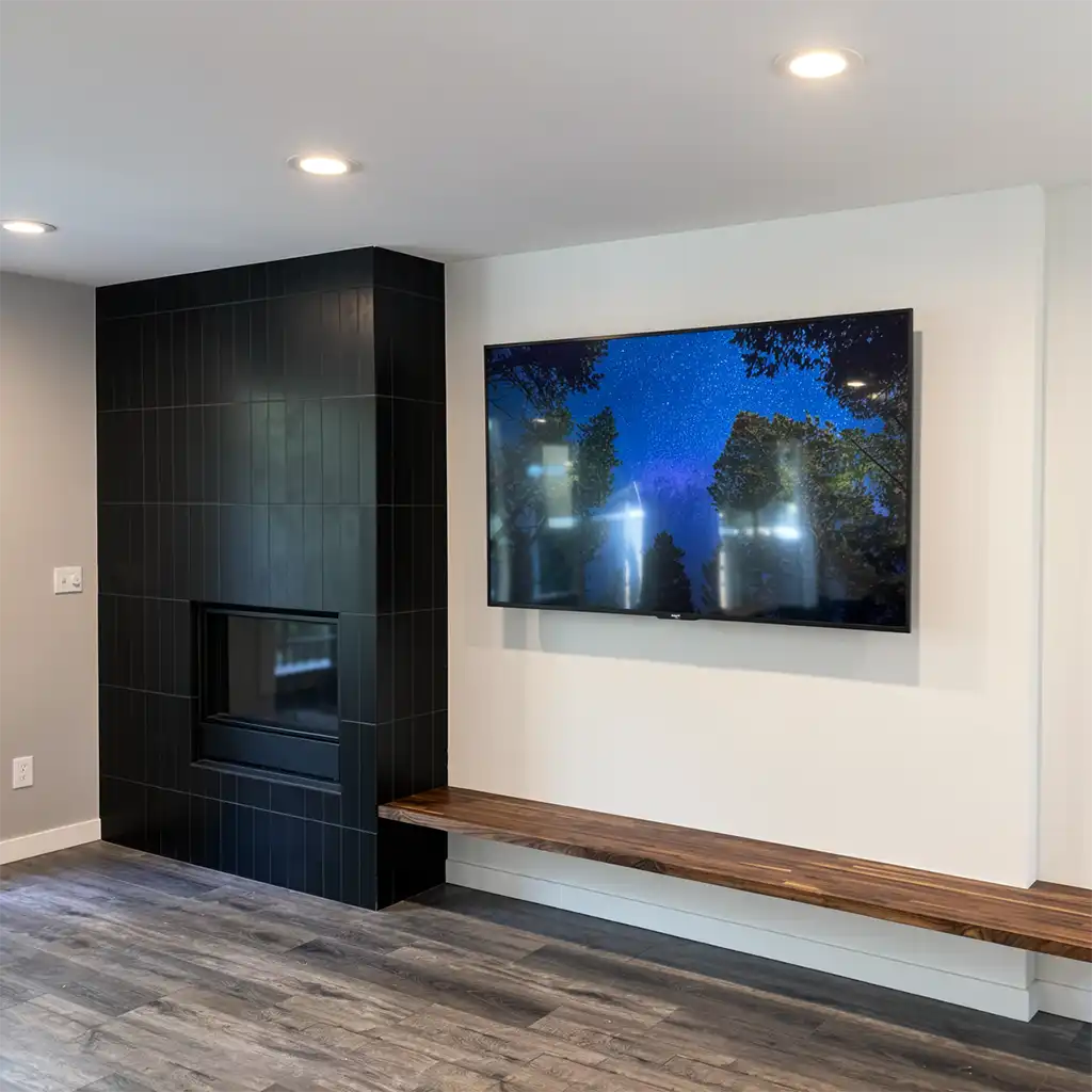 modern minimalist living room interior featuring a sleek black tiled fireplace with vertical paneling and a recessed glass-front firebox, positioned to the left; a large flat-screen television mounted on a pristine white wall displaying a vivid night sky with stars and tree silhouettes, centered above a low-profile wooden bench with natural grain finish and white base, all set against light gray walls and dark gray wood-plank flooring under recessed ceiling spotlights, captured in a wide-angle shot from eye level with shallow depth of field emphasizing the TV and fireplace as primary focal points