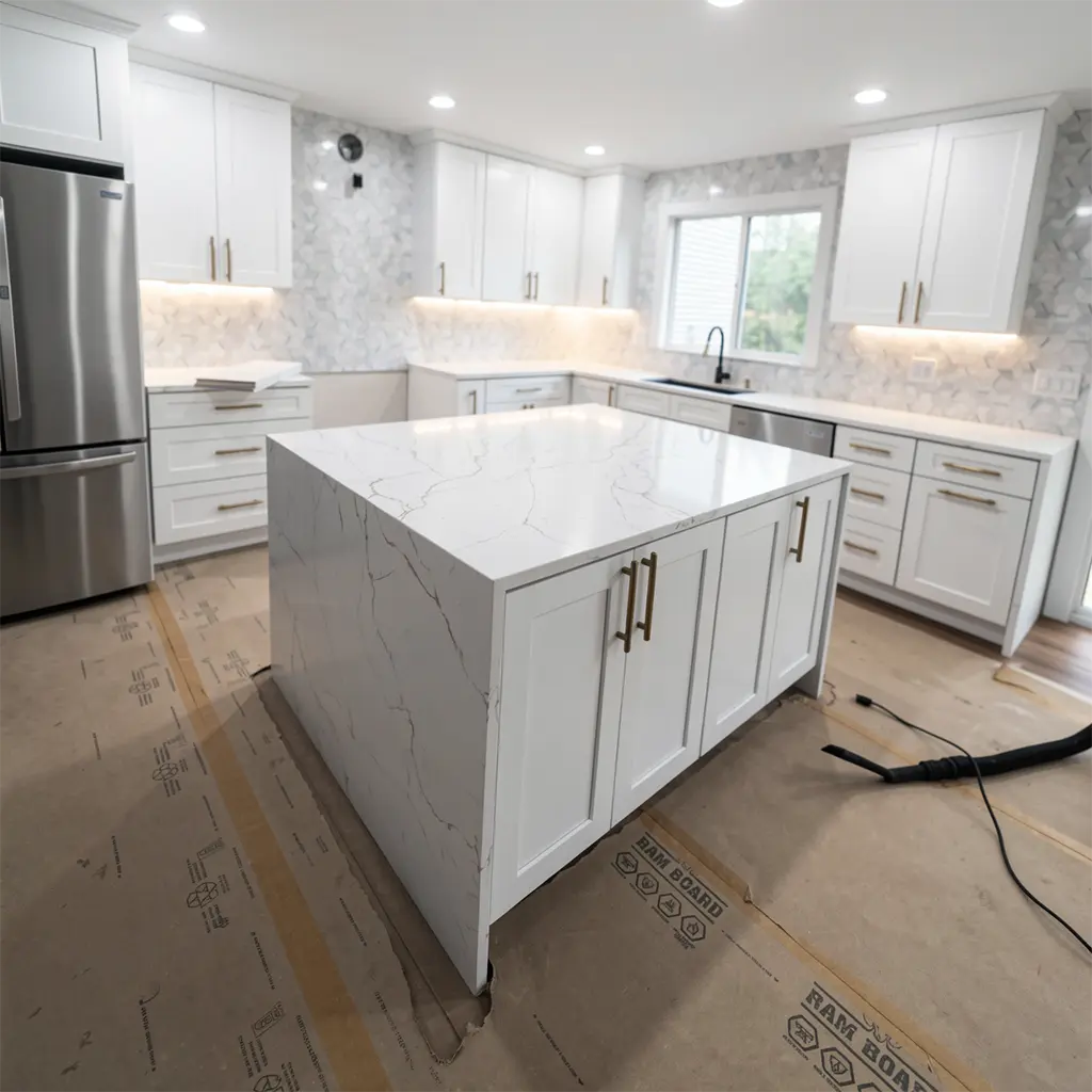 modern minimalist kitchen interior featuring white shaker-style cabinetry with gold-toned hardware, a central island with white marble countertops displaying natural veining, stainless steel refrigerator and dishwasher, under-cabinet LED lighting, light gray mosaic tile backsplash, recessed ceiling lights, wooden floor partially covered with protective RAM BOARD sheeting, black vacuum hose on floor, window with blinds showing greenery outside, no people present
