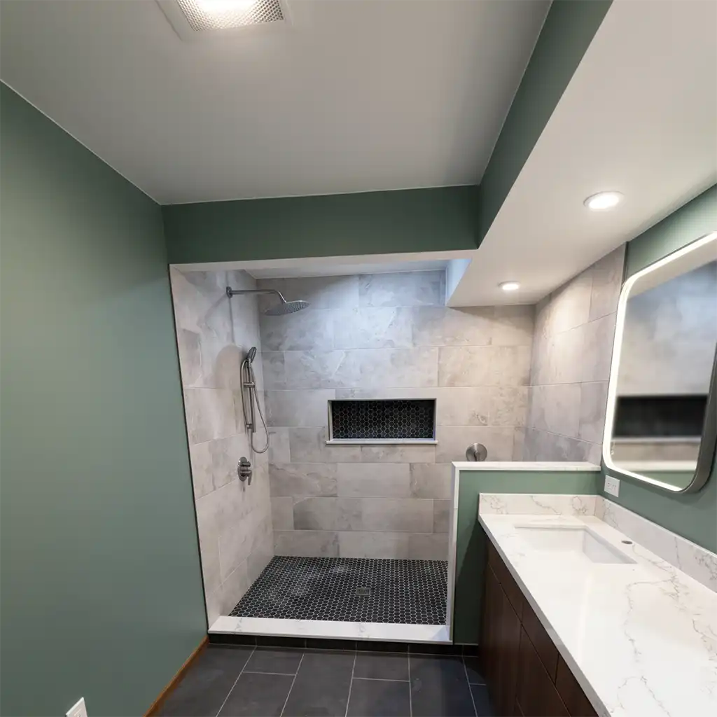 modern minimalist bathroom interior featuring a walk-in shower with light gray marble tiles, black hexagonal floor tiles, and a recessed niche with dark hexagonal tiling; vanity with white marble countertop and dark wood cabinetry, framed by sage green walls and ceiling trim; recessed LED lighting and a rectangular mirror with integrated backlighting mounted above the sink; overhead ventilation grille visible in ceiling; no people present