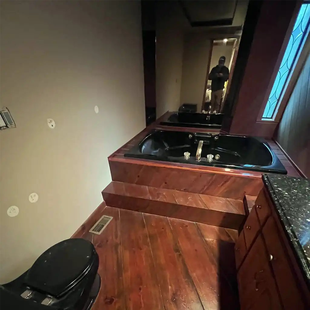 a dimly lit, opulent bathroom interior featuring a large black jacuzzi tub with chrome fixtures embedded in a raised wooden platform, surrounded by rich dark wood cabinetry and flooring, with a black toilet to the left and a stained glass window casting blue light on the right wall; camera capturing the scene with shallow depth of field and warm artificial lighting accentuating the wood grain and glossy surfaces
