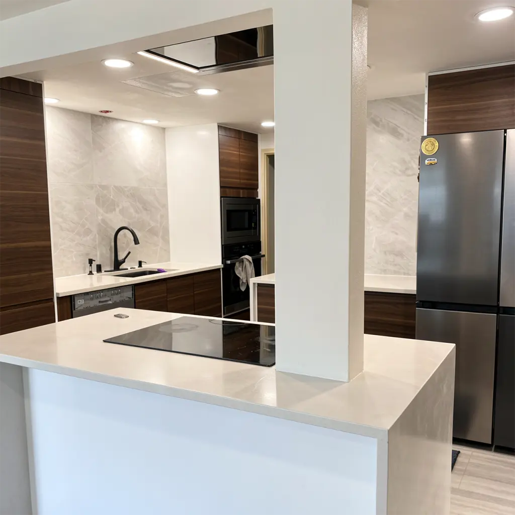 modern minimalist kitchen interior featuring sleek white quartz countertops, dark wood cabinetry with horizontal grain texture, stainless steel appliances including a large refrigerator with decorative magnets, an induction cooktop embedded in the island, black high-arc faucet over a sink, recessed ceiling lights casting even illumination, marble-patterned backsplash tiles, a central white structural pillar dividing the space, light wood flooring, and a clean, uncluttered composition with shallow depth of field emphasizing the foreground island