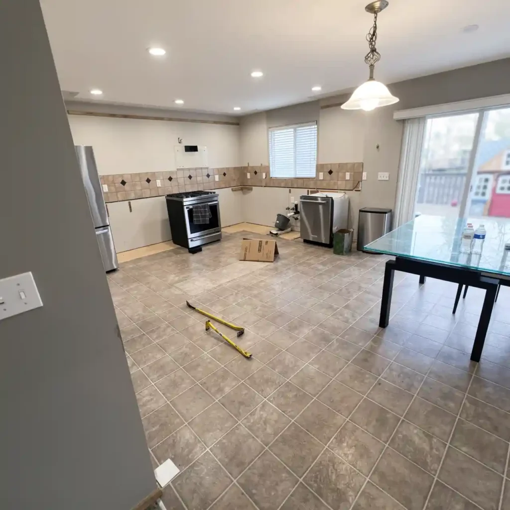 photorealistic interior shot of a modern kitchen and dining area under construction with beige tiled flooring, stainless steel appliances including refrigerator, stove, and dishwasher, unfinished light gray walls with partial tile backsplash featuring diamond-patterned accent tiles, recessed ceiling lights and one ornate hanging pendant lamp, glass-top dining table with black legs positioned near sliding glass doors revealing an outdoor yard with a red playhouse in the background, scattered tools including yellow measuring tape on floor, cardboard box, trash bins, and empty water bottles on table