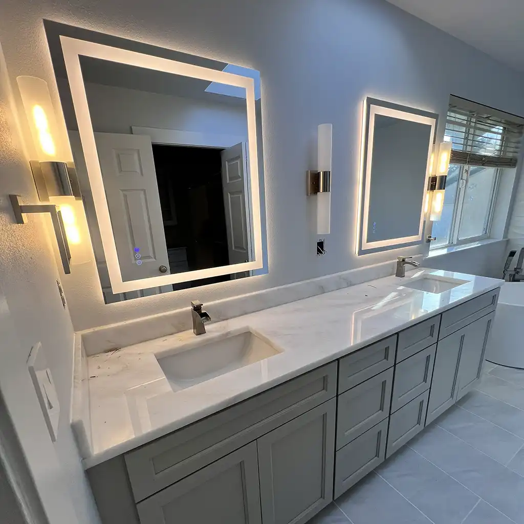 modern minimalist bathroom interior featuring two large rectangular LED-framed mirrors with integrated lighting, mounted above a long white marble countertop with dual undermount sinks and brushed nickel faucets; light gray shaker-style cabinetry with recessed paneling, flanked by matching cylindrical wall sconces emitting warm ambient glow; floor covered in large-format matte gray tiles, with a window to the right showing blinds and exterior view, and a freestanding white bathtub partially visible on the far right; walls painted in soft cool blue-gray tone, ceiling with recessed lighting