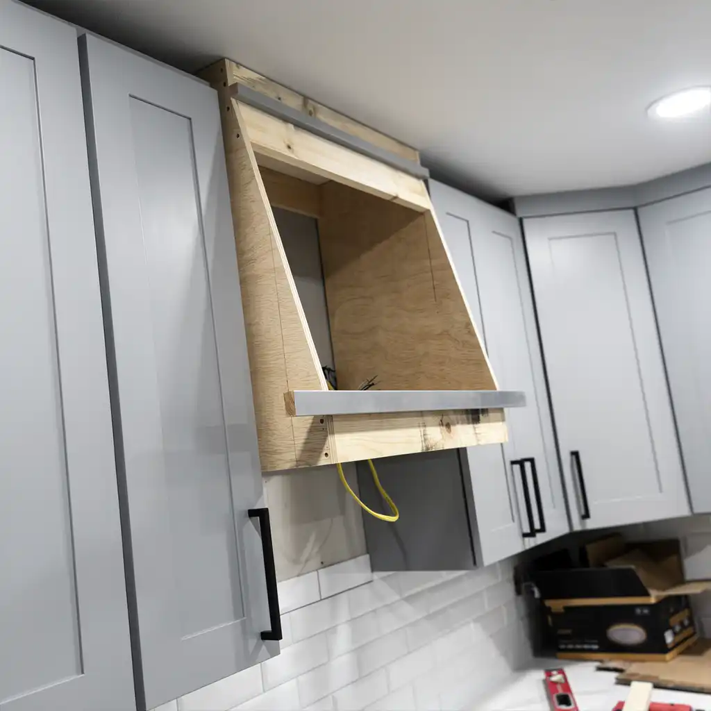 photorealistic interior shot of a modern kitchen under construction featuring light gray shaker-style cabinets with black handles, a partially assembled wooden range hood with exposed plywood panels and a metallic trim, yellow electrical wiring hanging from the underside, white subway tile backsplash, and scattered construction materials including cardboard boxes and tools on the countertop; overhead recessed lighting illuminates the scene from above with soft, even illumination
